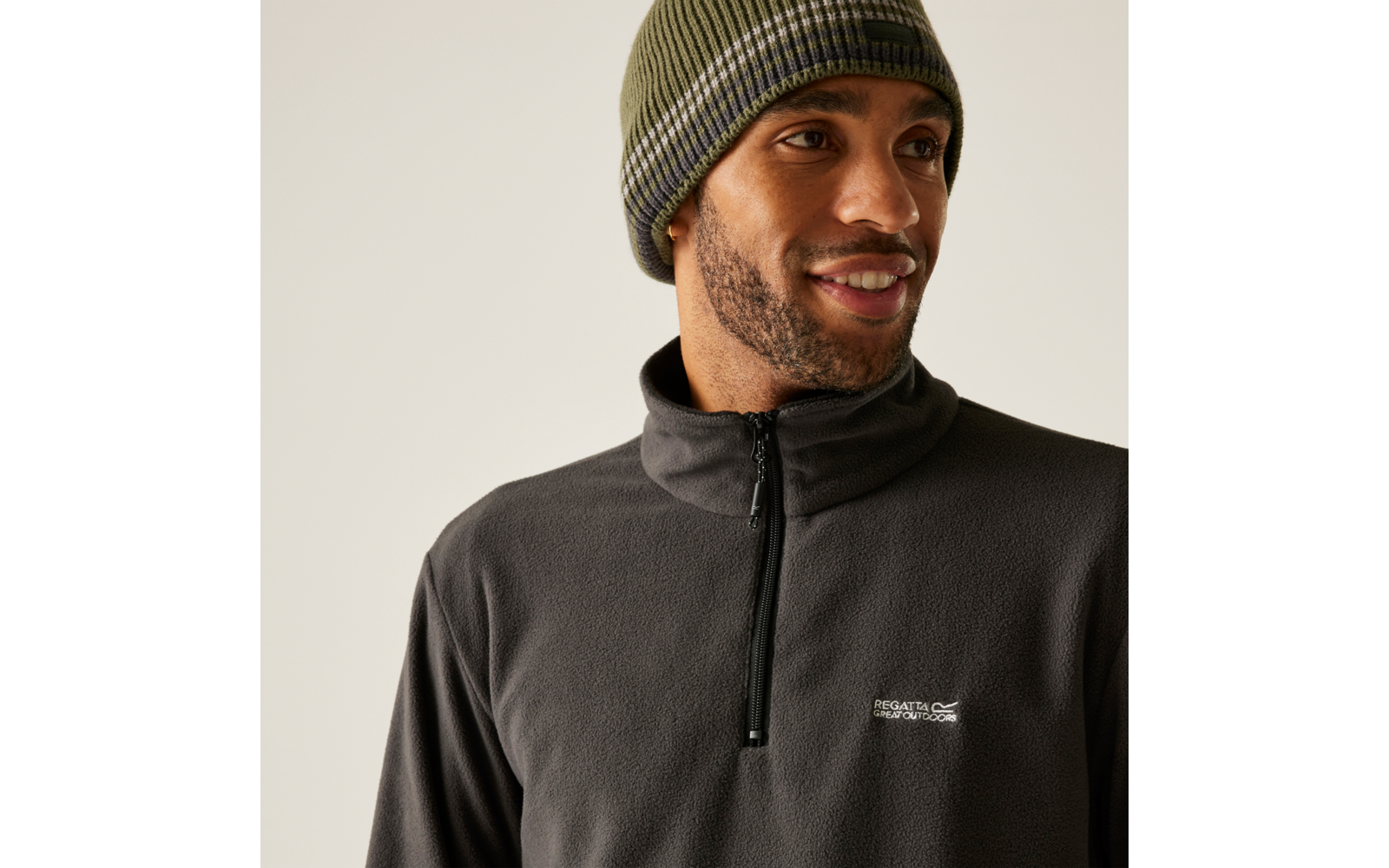 Personne avec bonnet en maille olive et polaire gris foncé à demi-fermeture éclair, souriante, regard vers la droite