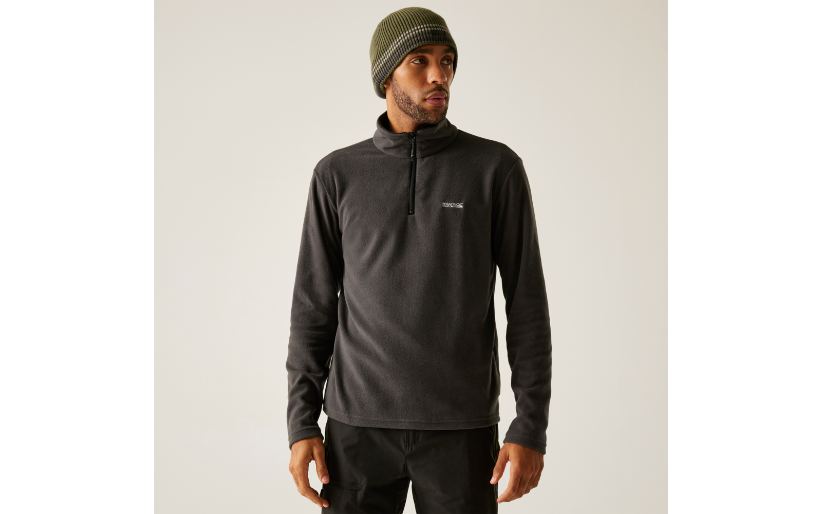 Personne portant une polaire gris foncé à demi-fermeture éclair, un bonnet tricoté vert olive et un pantalon noir sur fond clair