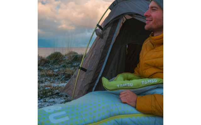 Person sitting in the entrance of a tent, wearing a yellow jacket and holding an inflatable pillow labeled 'TAMBU'; spotted inflatable mattress in the foreground, frosty grass and a lake under a cloudy sky in the background