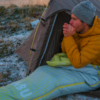 Person in yellow puffer jacket and gray beanie sits at a tent entrance with hands near the mouth; sleeping bag and lightly snow-covered ground visible
