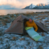 Person in a yellow jacket and gray beanie sits at the entrance of a brown tent, partially in a light green-blue sleeping bag; snow-dusted grass; sea and snow-covered mountains in the background