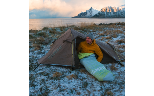 Person in a yellow jacket and gray beanie sits at the entrance of a brown tent, partially in a light green-blue sleeping bag; snow-dusted grass; sea and snow-covered mountains in the background