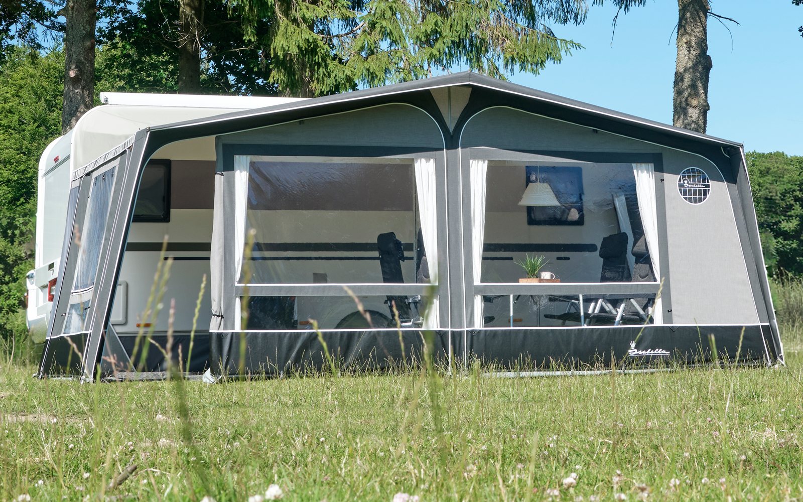 Weißer Wohnwagen mit angebautem Vorzelt auf grüner Wiese, im Hintergrund hohe Nadelbäume und blauer Himmel