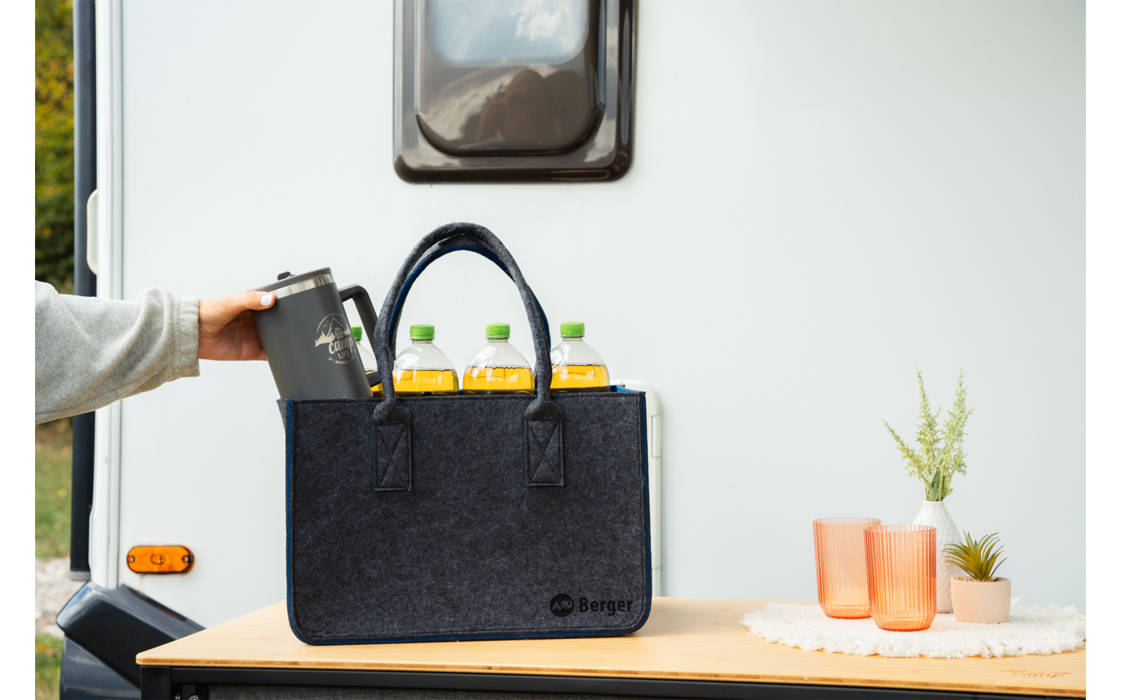 Schwarze Filztasche auf Holztisch vor weißer Wohnwagenwand; drei Flaschen mit gelber Flüssigkeit, eine Hand stellt eine Thermoskanne hinein; daneben zwei orange Becher und eine kleine Topfpflanze