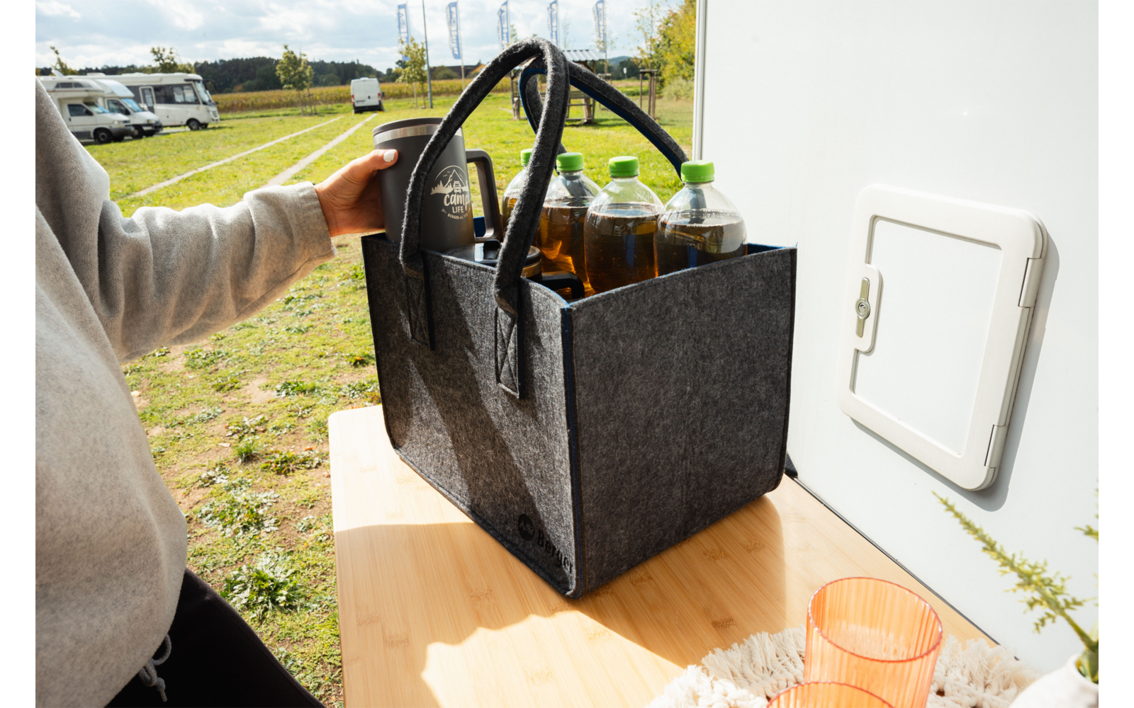 Graue Filztragetasche mit zwei Griffen auf einem Holztisch neben einer weißen Wohnwagenaußentür; im Inneren vier Flaschen mit grünen Verschlüssen; eine Hand in hellgrauem Sweatshirt hält die Tasche; im Hintergrund Rasenfläche und geparkte Wohnmobile