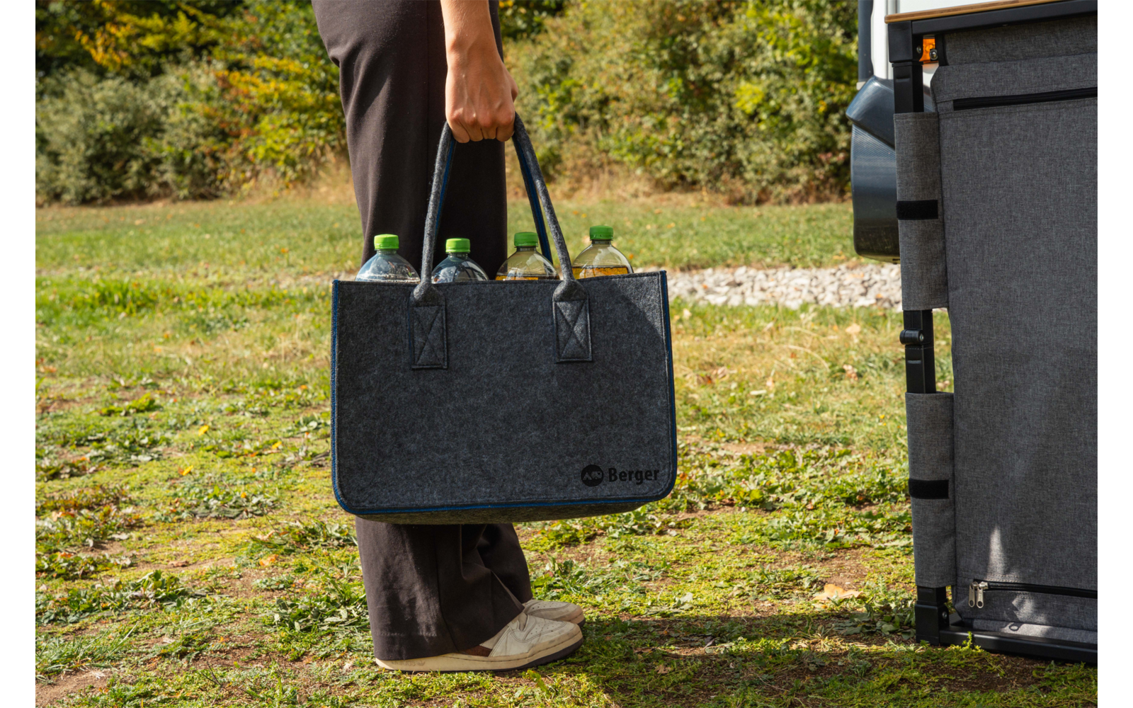 Hand einer Person hält eine graue Filztasche mit mehreren Flaschen mit grünen Verschlüssen; Beine und Schuhe sichtbar, Person steht auf Gras neben einem Fahrzeug