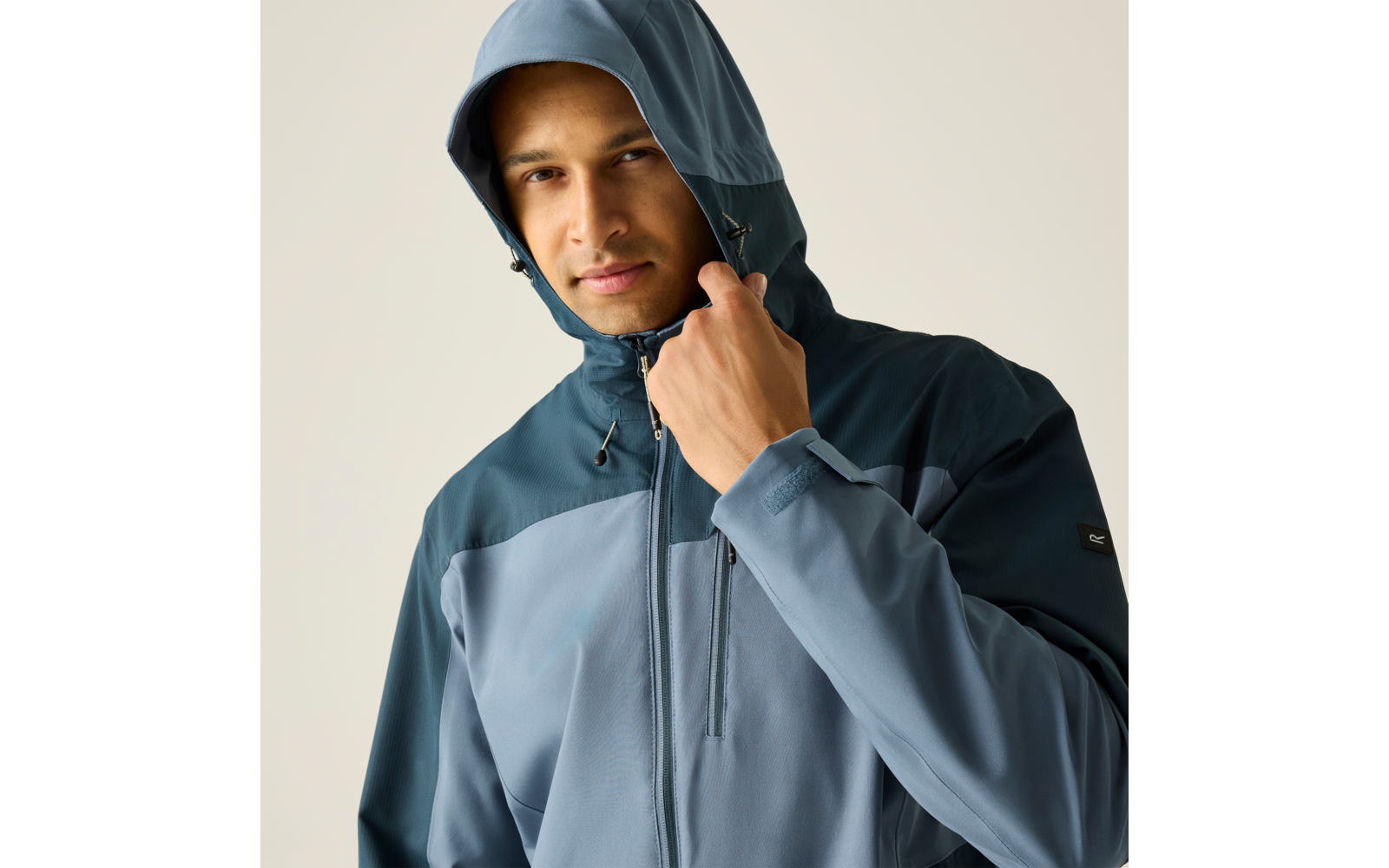 Person trägt eine blau-grüne Kapuzenjacke und hält die Kapuze mit der rechten Hand; heller einfarbiger Hintergrund