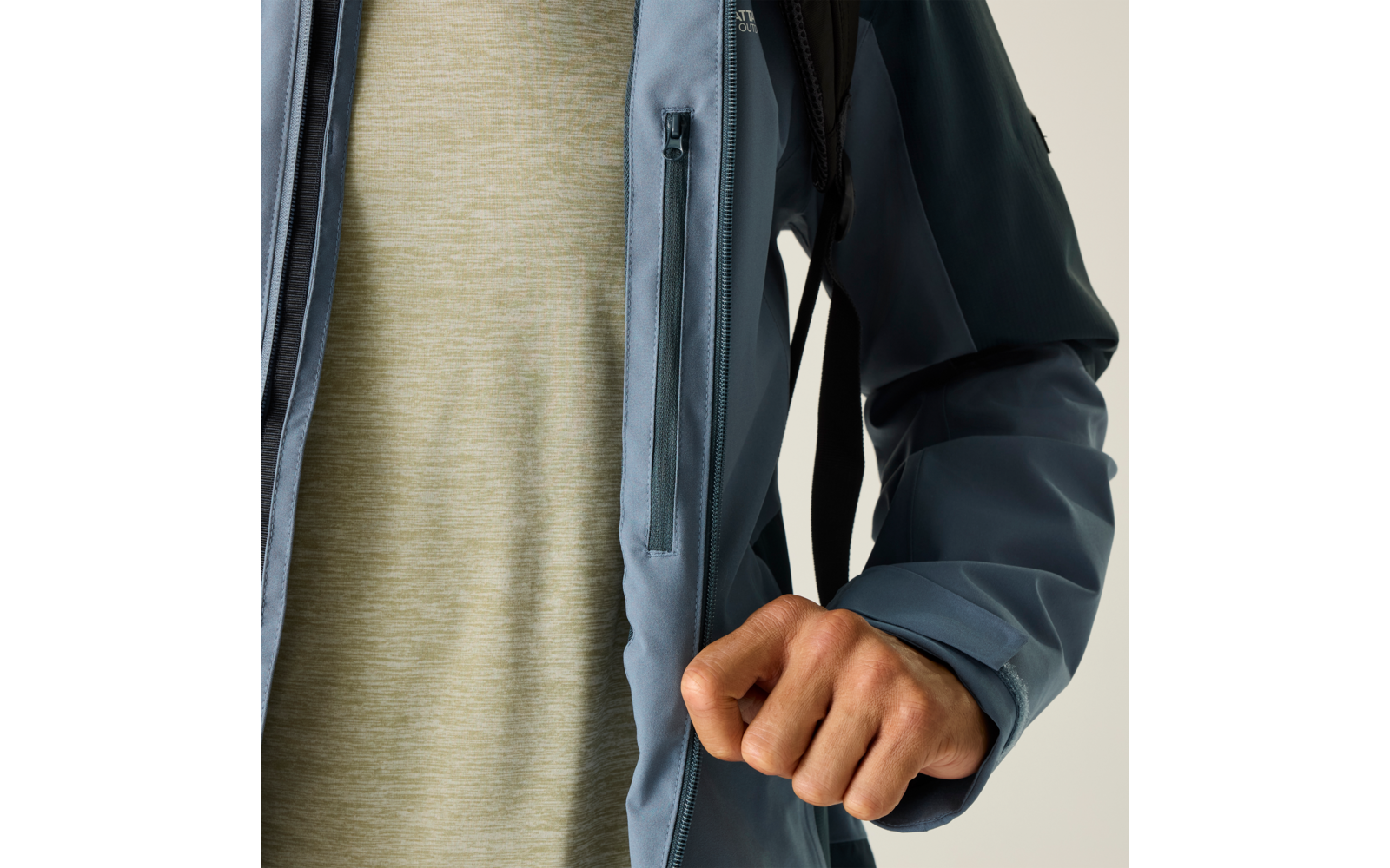 Oberkörper einer Person in blauer Jacke mit Reißverschlusstasche, hellgrünes meliertes T‑Shirt darunter, rechte Hand zur Faust vor dem Körper, schwarzer Rucksackriemen, heller Hintergrund