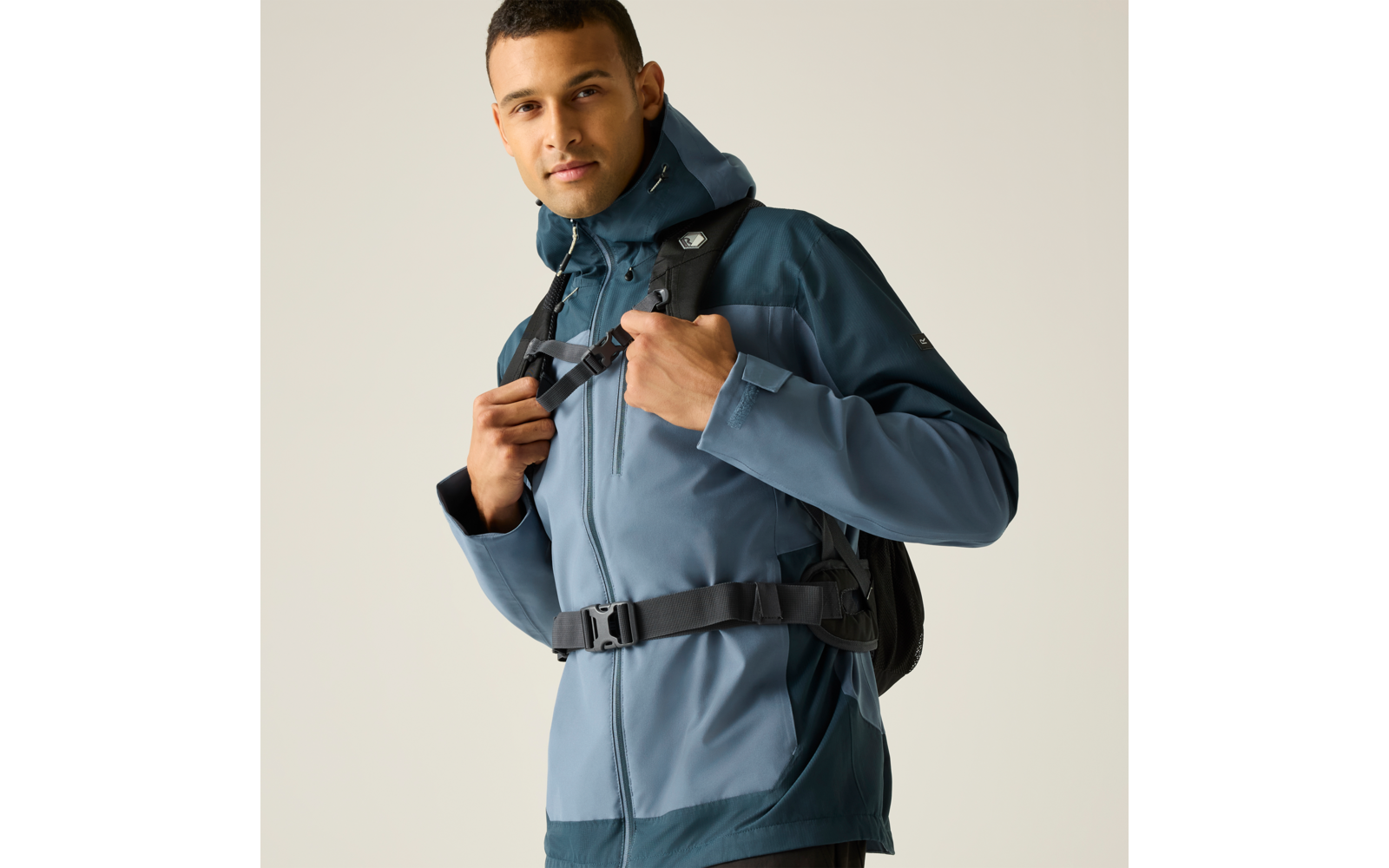 Person mit blauer Outdoorjacke und aufgesetzter Kapuze, trägt einen schwarzen Rucksack mit Brustgurt und hält den Träger