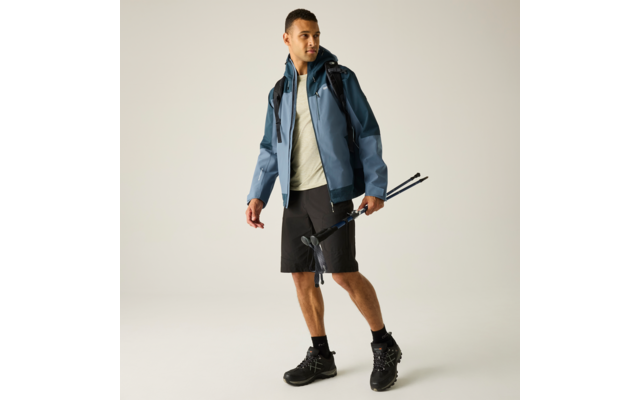 Mann in blauer Jacke, helles T‑Shirt, schwarze Shorts, Wanderschuhe, Rucksack, hält zwei Trekkingstöcke, heller Hintergrund
