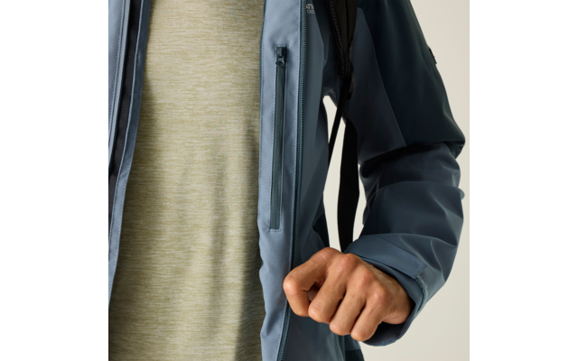 Oberkörper einer Person in blauer Jacke mit Reißverschlusstasche, hellgrünes meliertes T‑Shirt darunter, rechte Hand zur Faust vor dem Körper, schwarzer Rucksackriemen, heller Hintergrund