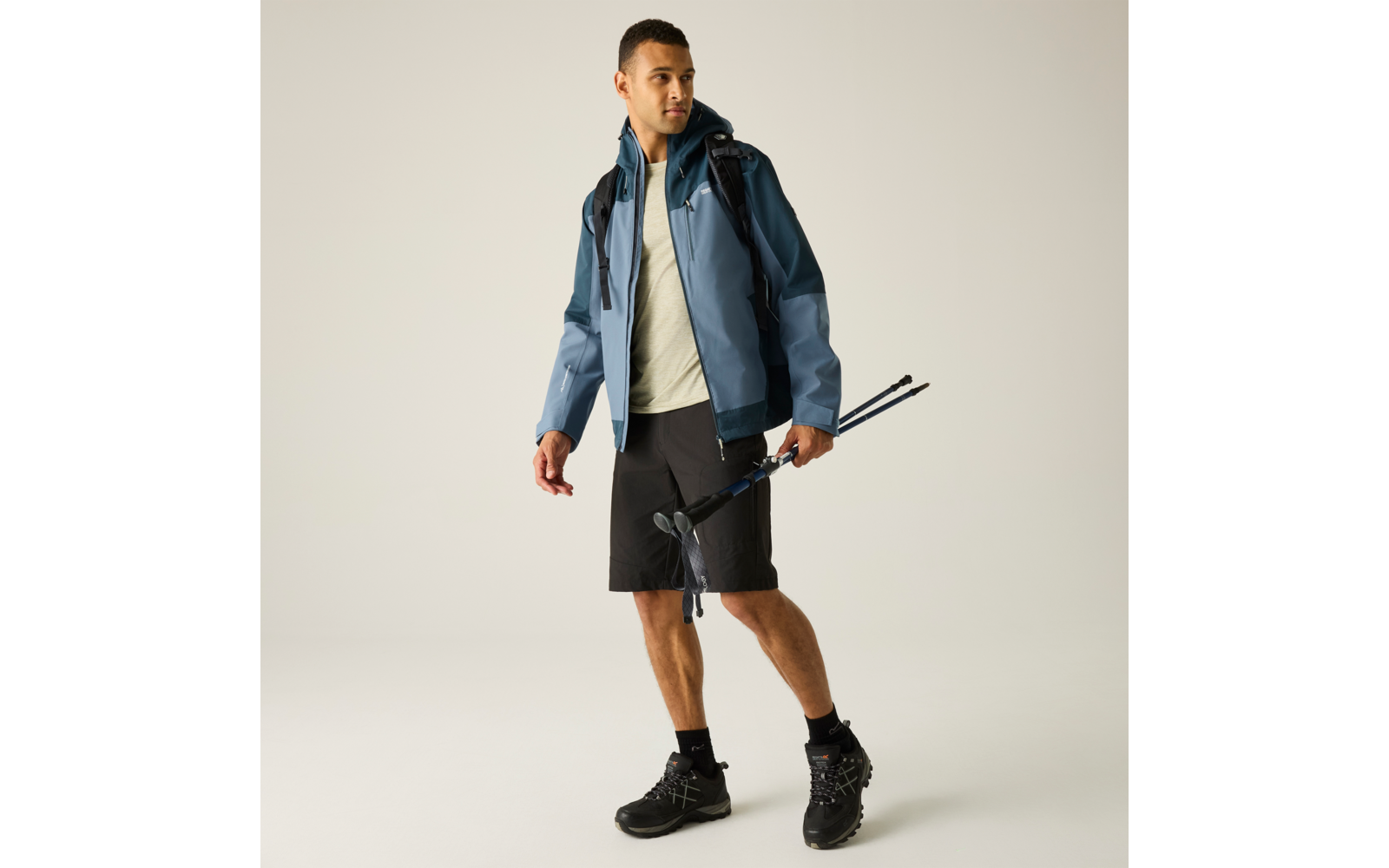 Mann in blauer Jacke, helles T‑Shirt, schwarze Shorts, Wanderschuhe, Rucksack, hält zwei Trekkingstöcke, heller Hintergrund
