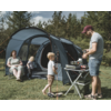 Man at a portable stove on a folding table in front of a large open tent, two children nearby, a woman seated on a camping chair; grass and trees in the background