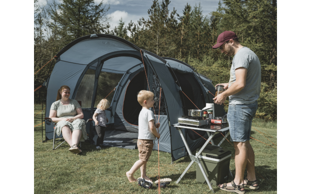 Man at a portable stove on a folding table in front of a large open tent, two children nearby, a woman seated on a camping chair; grass and trees in the background