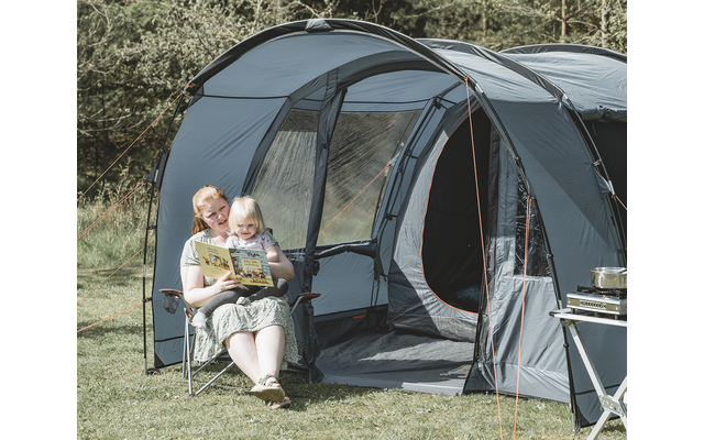 An adult and a child sit on a folding chair in front of a large gray tent; the adult holds an open book, the child sits on their lap; grass and trees in the background; a camping table with a stove is at the right