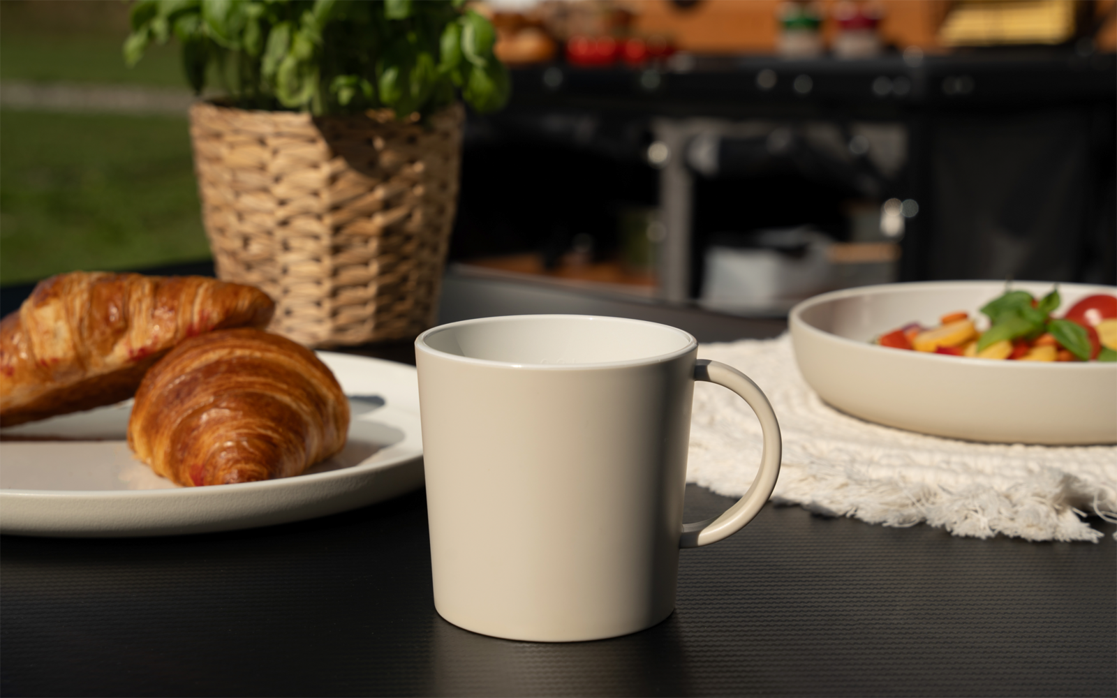 Beige Keramiktasse auf dunklem Tisch; links zwei Croissants auf einem Teller; rechts Schüssel mit Obstsalat; im Hintergrund geflochtener Topf mit grüner Pflanze; unscharfer Außenhintergrund