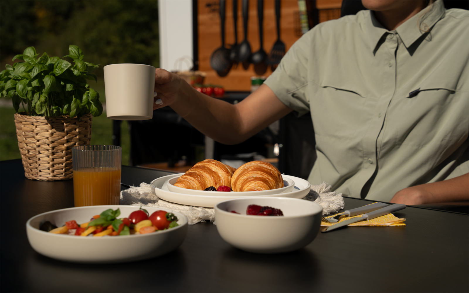 Dunkler Tisch mit weißem Teller mit zwei Croissants, zwei weißen Schüsseln (Salat mit Cherrytomaten; rote Beeren), Glas Orangensaft und geflochtener Topf mit Basilikum. Teilanschnitt einer Person im hellgrünen Hemd, Oberkörper und Arm sichtbar, hält 