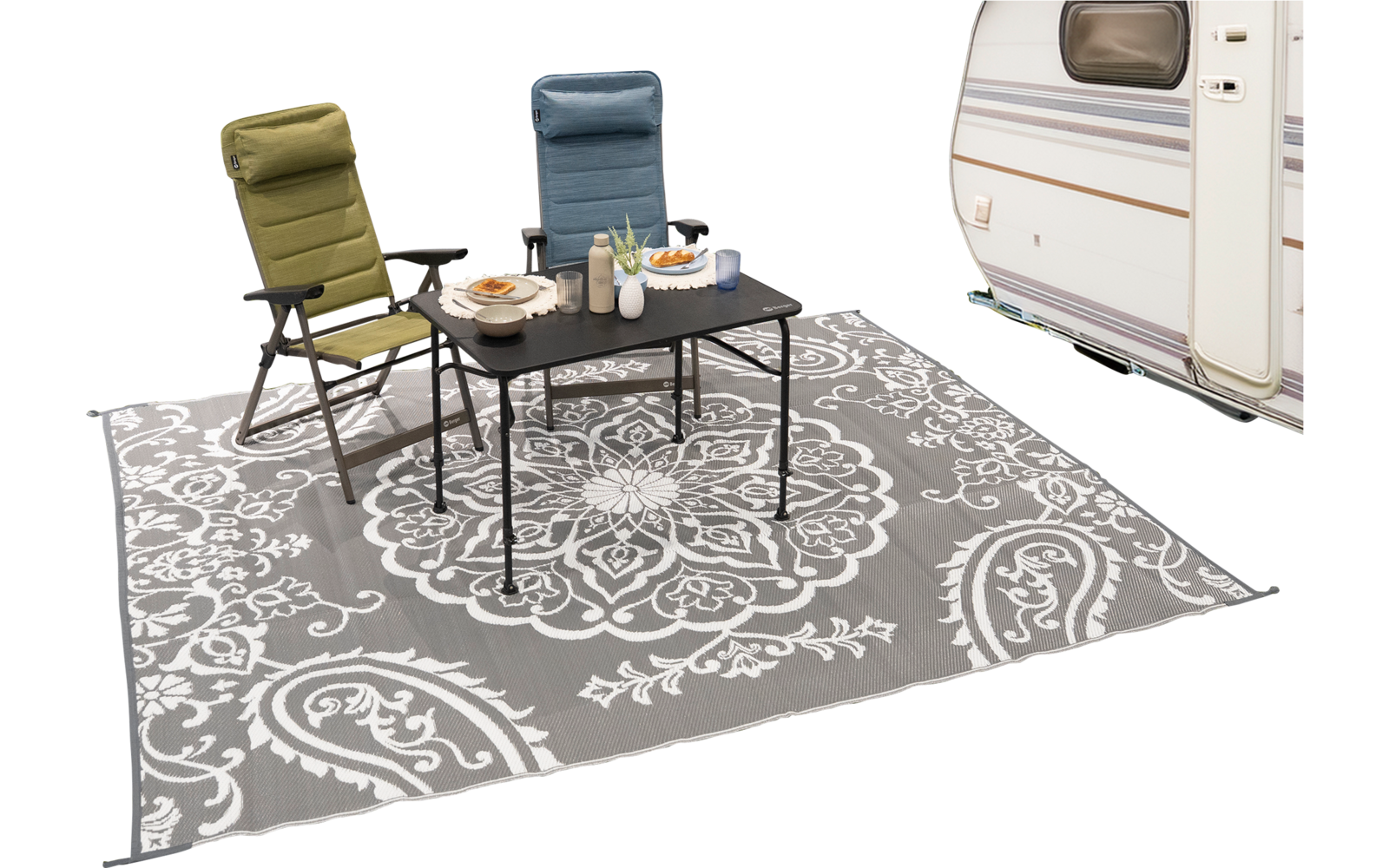 Zwei Campingstühle (grün, blau) und ein schwarzer Klapptisch mit Tellern, Flaschen und Schalen auf einem grauen Teppich mit weißem Ornamentmuster; rechts teilweise ein Wohnwagen sichtbar