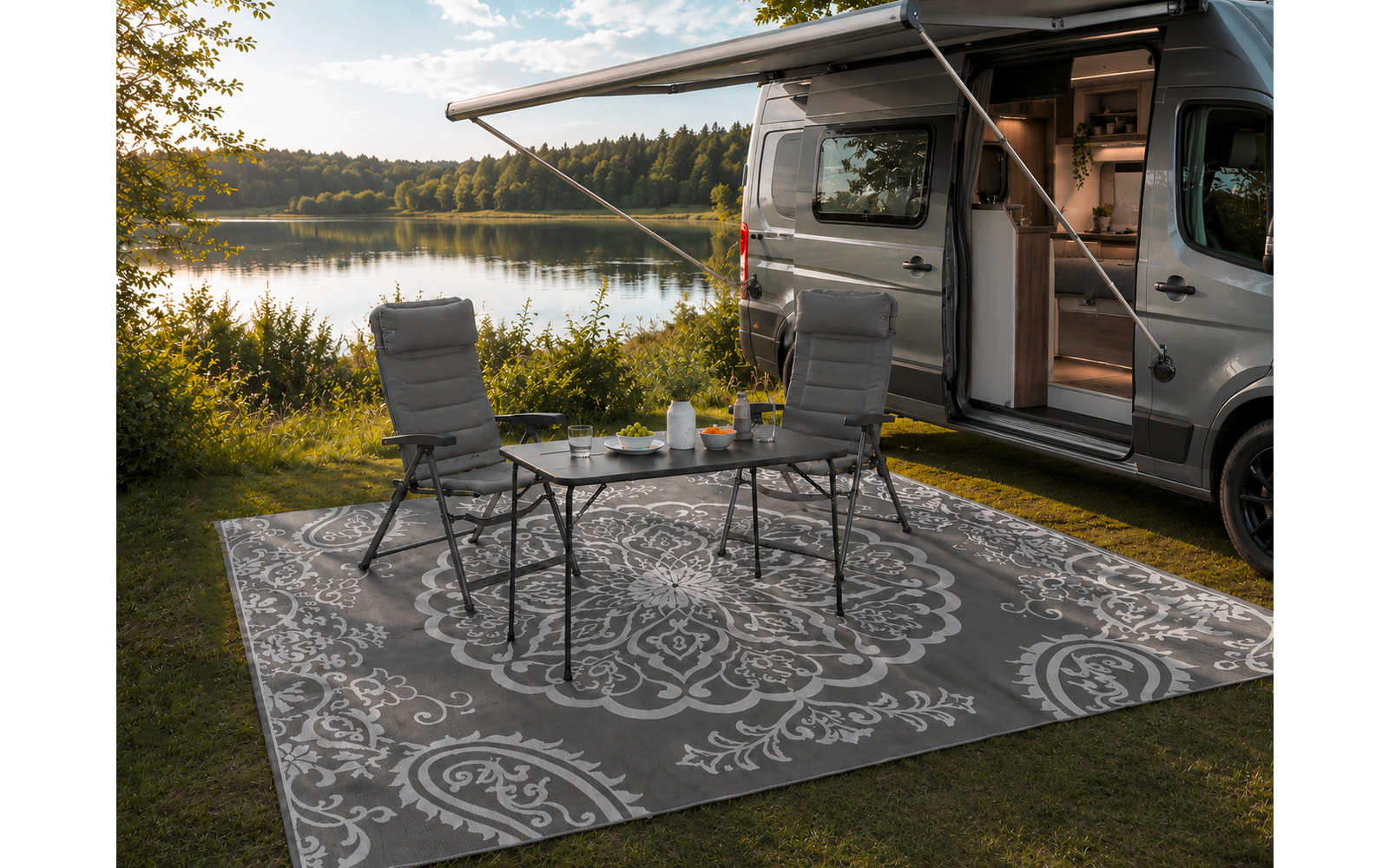 Furgoneta camper gris con puerta lateral abierta y toldo desplegado; delante una alfombra exterior con estampado, dos sillas de camping plegables y una mesita con una jarra y vasos, en la orilla de un lago con árboles al fondo