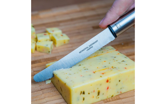 Couteau dentelé portant l'inscription 'MONOPOL GERMANY rostfrei' coupe un bloc de fromage jaune avec des morceaux verts et rouges sur une planche en bois ; dés de fromage en arrière-plan