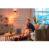 Two people in a living room: one sits on the floor in front of a sofa, the other kneels behind the sofa with hands on the first person's shoulders. Visible: coffee table with potted plant, large houseplant by the window, lamp, framed wall art, and a portable heater on the floor