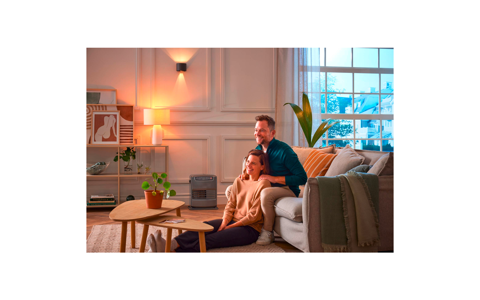 Two people in a living room: one sits on the floor in front of a sofa, the other kneels behind the sofa with hands on the first person's shoulders. Visible: coffee table with potted plant, large houseplant by the window, lamp, framed wall art, and a portable heater on the floor