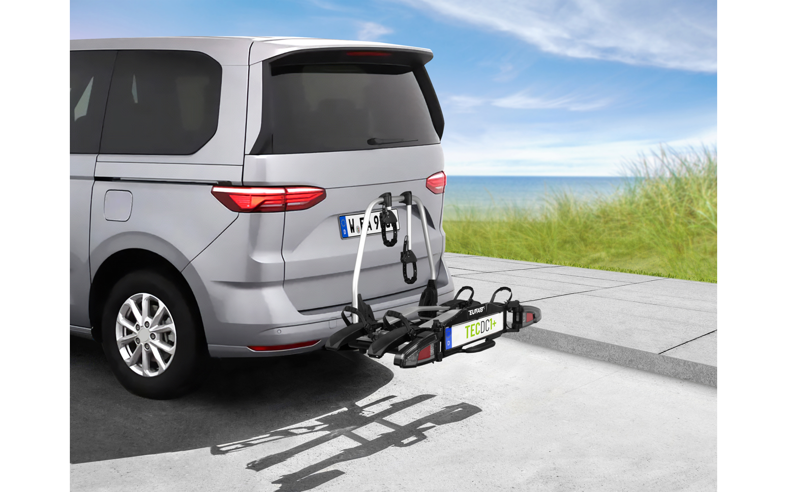 Heck eines silbernen Vans mit an der Anhängerkupplung montiertem Fahrradträger mit zwei Fahrrädern, Parkfläche, Grasdüne und Himmel im Hintergrund