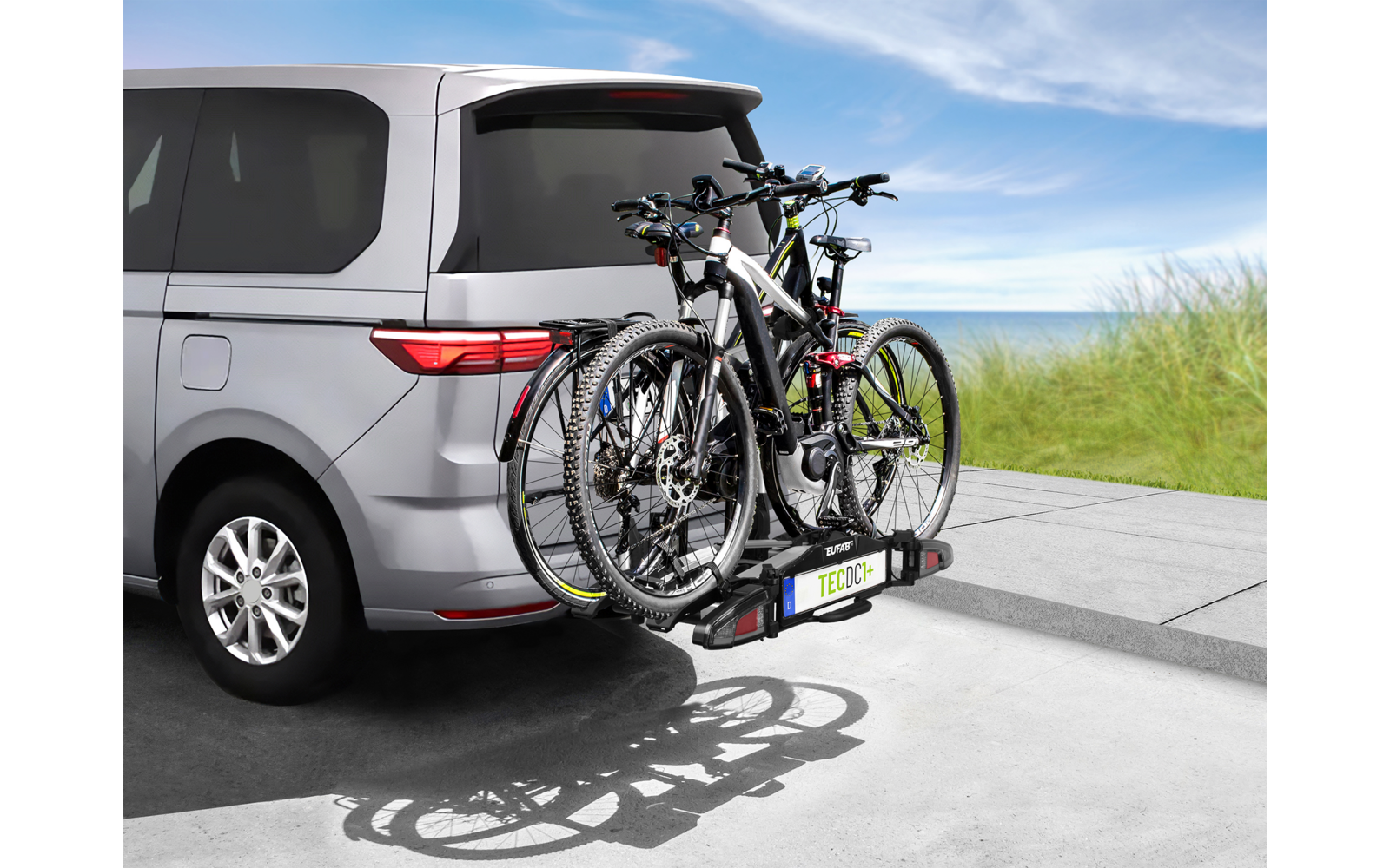 Silberner Van von hinten links, an der Heckklappe montierter Fahrradträger mit drei Fahrrädern, Betonfläche, Grasdüne, Meer und blauem Himmel im Hintergrund