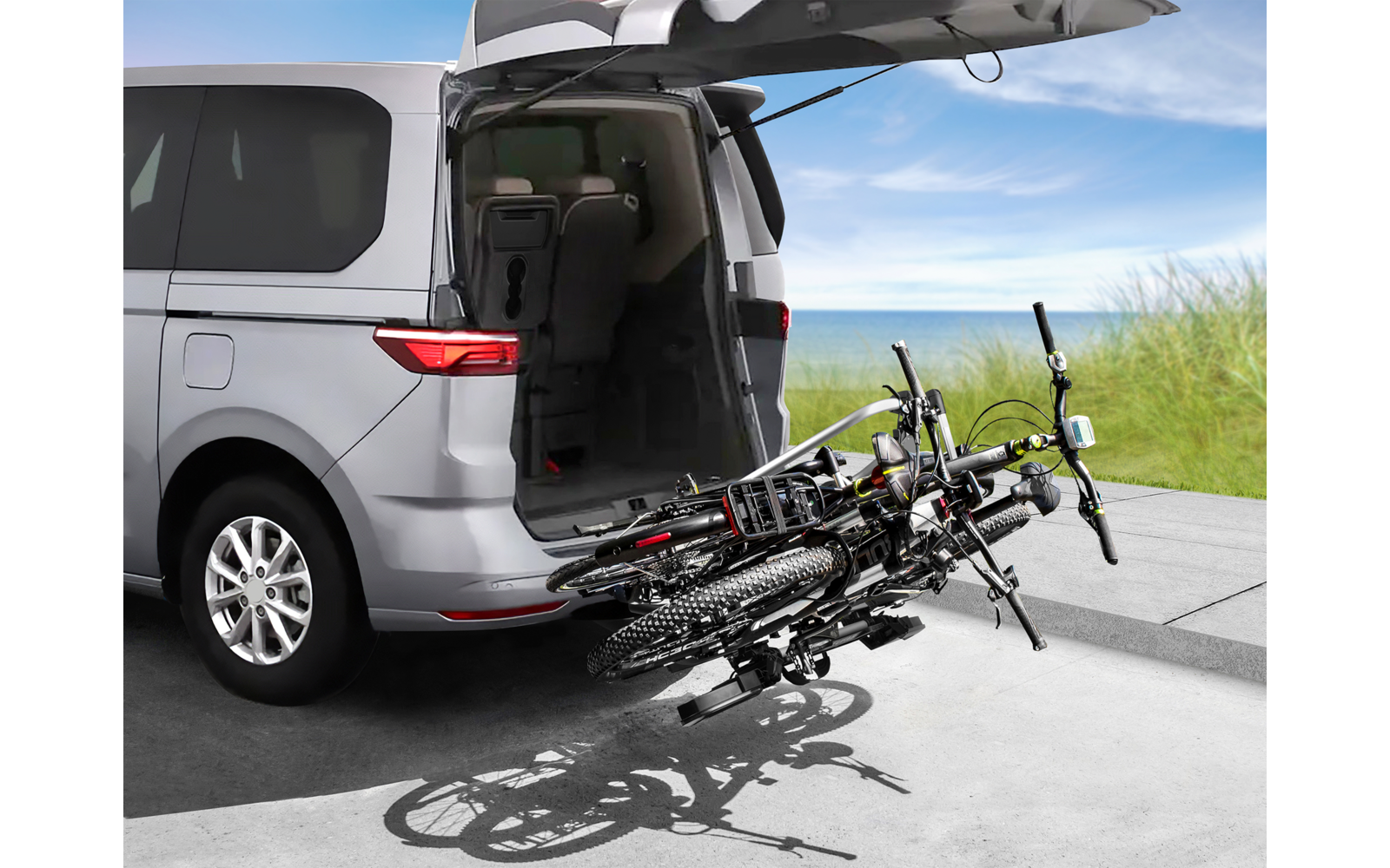Silberner Van mit geöffneter Heckklappe und zwei Fahrrädern an einem Heckträger, geparkt auf einem Weg neben Gras und blauem Himmel