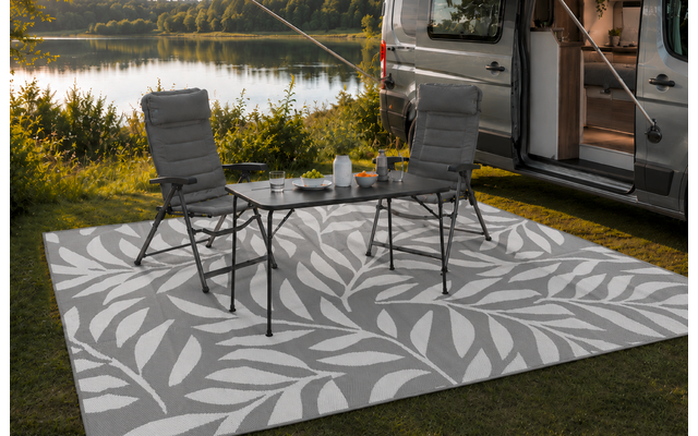 Camper geparkeerd aan de oever van een meer, schuifdeur open. Twee campingstoelen en een klein inklapbaar tafeltje op een licht patroon buitenkleed. Theepot en kopjes op de tafel. Gras, bomen en meer op de achtergrond, bewolkte lucht