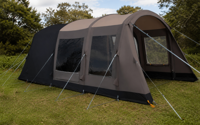 Grote grijze tent met drie rechthoekige ramen, open ingang, scheerlijnen en stokken, op groen gras, bomen en lucht op de achtergrond