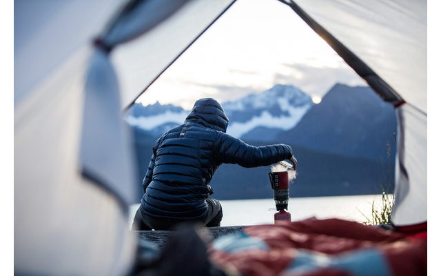 Veduta da una tenda aperta: persona con piumino scuro seduta che tiene un fornello da campeggio; lago e montagne sullo sfondo