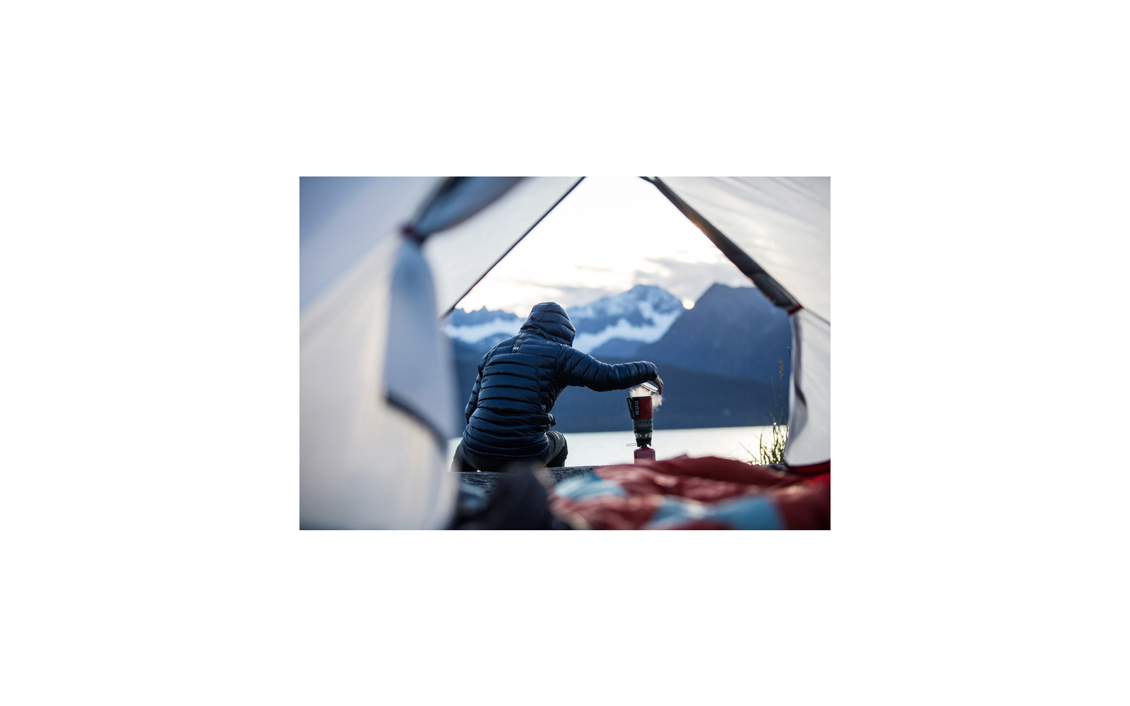 Veduta da una tenda aperta: persona con piumino scuro seduta che tiene un fornello da campeggio; lago e montagne sullo sfondo