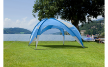 Blauer Pavillon auf grünem Rasen am Seeufer; im Hintergrund See, Boote, Personen am Steg, Bäume und Hügel unter leicht bewölktem Himmel
