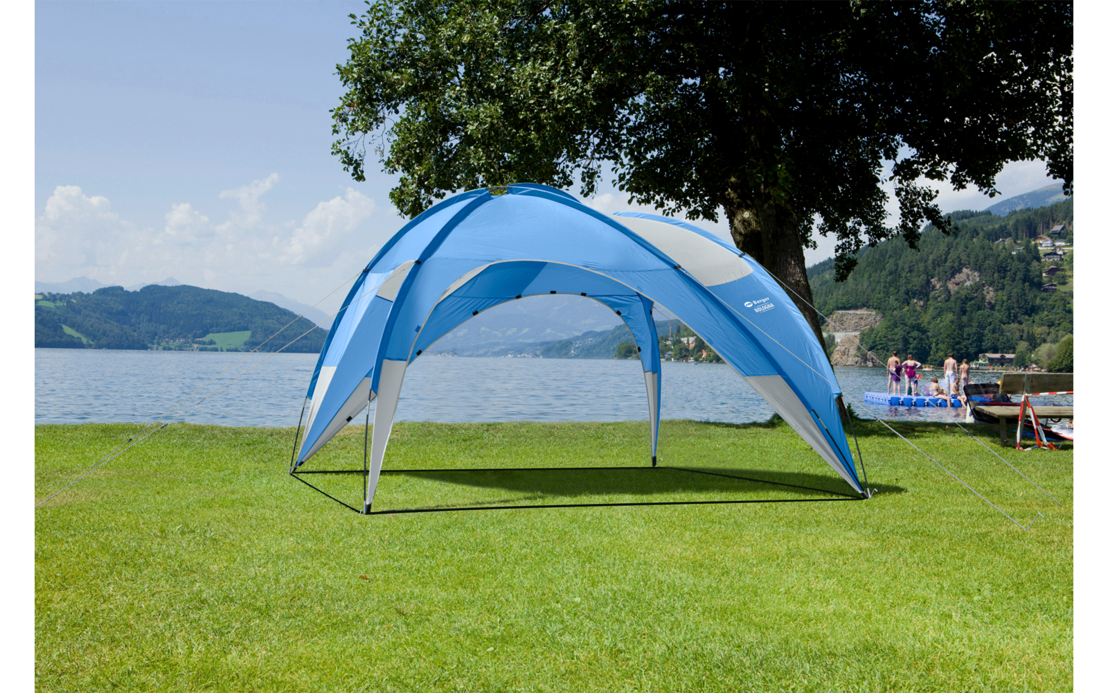 Blauer Pavillon auf grünem Rasen am Seeufer; im Hintergrund See, Boote, Personen am Steg, Bäume und Hügel unter leicht bewölktem Himmel