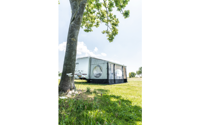 Weißer Wohnwagen mit grauem Vorzelt mit runden Fenstern auf einer Wiese, Baumstamm im Vordergrund, bewölkter Himmel