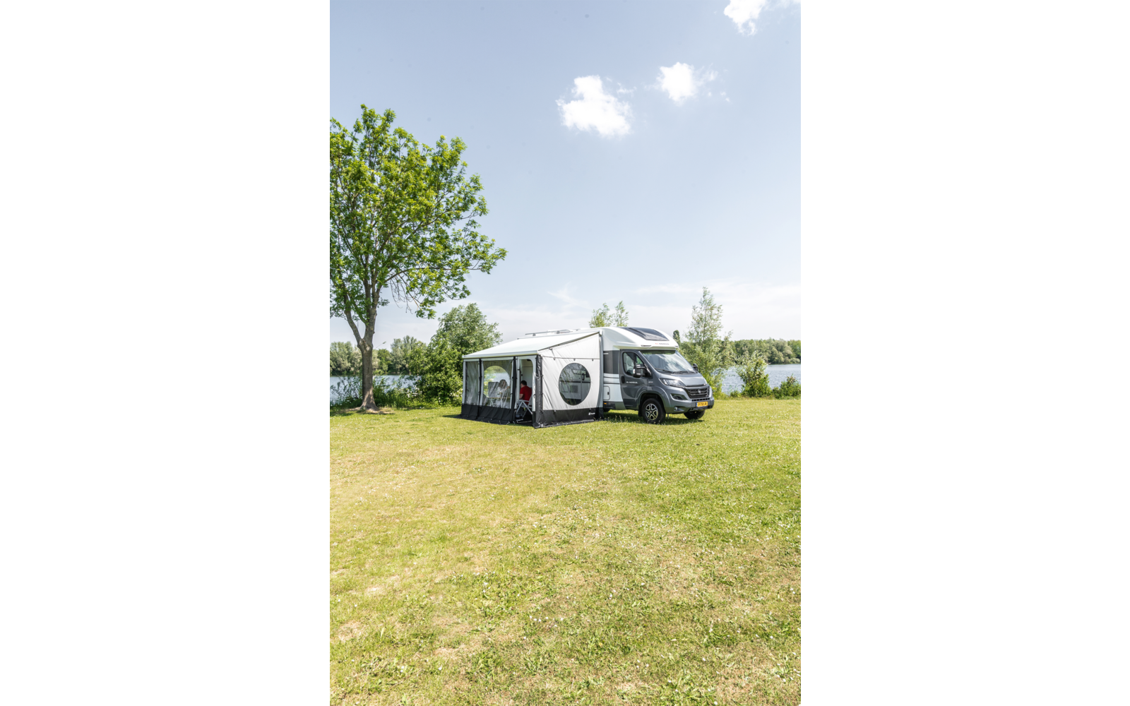 Wohnmobil mit angebautem Vorzelt auf einer Wiese neben einem See, Bäume und blauer Himmel mit vereinzelten Wolken