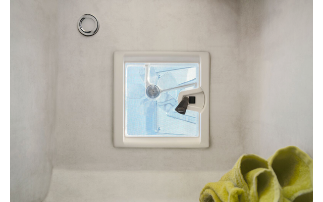 Quadratisches Lüftungsfenster mit weißem Rahmen und sichtbarem Propellerlüfter hinter blauer Abdeckung; schwarzer Griff rechts, runde Metallöffnung oben links, gefaltetes grünes Tuch unten rechts