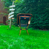Chaise pliante orange et noire sur gazon vert devant une haie ; à gauche un poteau en bois avec plusieurs flèches