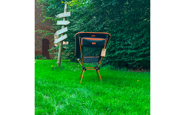 Chaise pliante orange et noire sur gazon vert devant une haie ; à gauche un poteau en bois avec plusieurs flèches