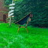 Chaise pliante noire avec armature orange sur herbe verte, panneau en bois et haie en arrière-plan