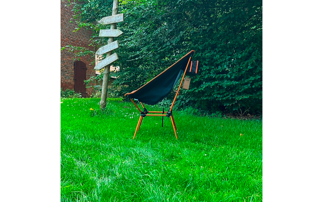 Chaise pliante noire avec armature orange sur herbe verte, panneau en bois et haie en arrière-plan