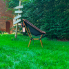 Chaise pliante noire avec armature orange sur herbe verte; à gauche un bâtiment en briques rouges et un poteau en bois avec plusieurs panneaux blancs; en arrière-plan une haie verte dense; une étiquette rose pend à la chaise
