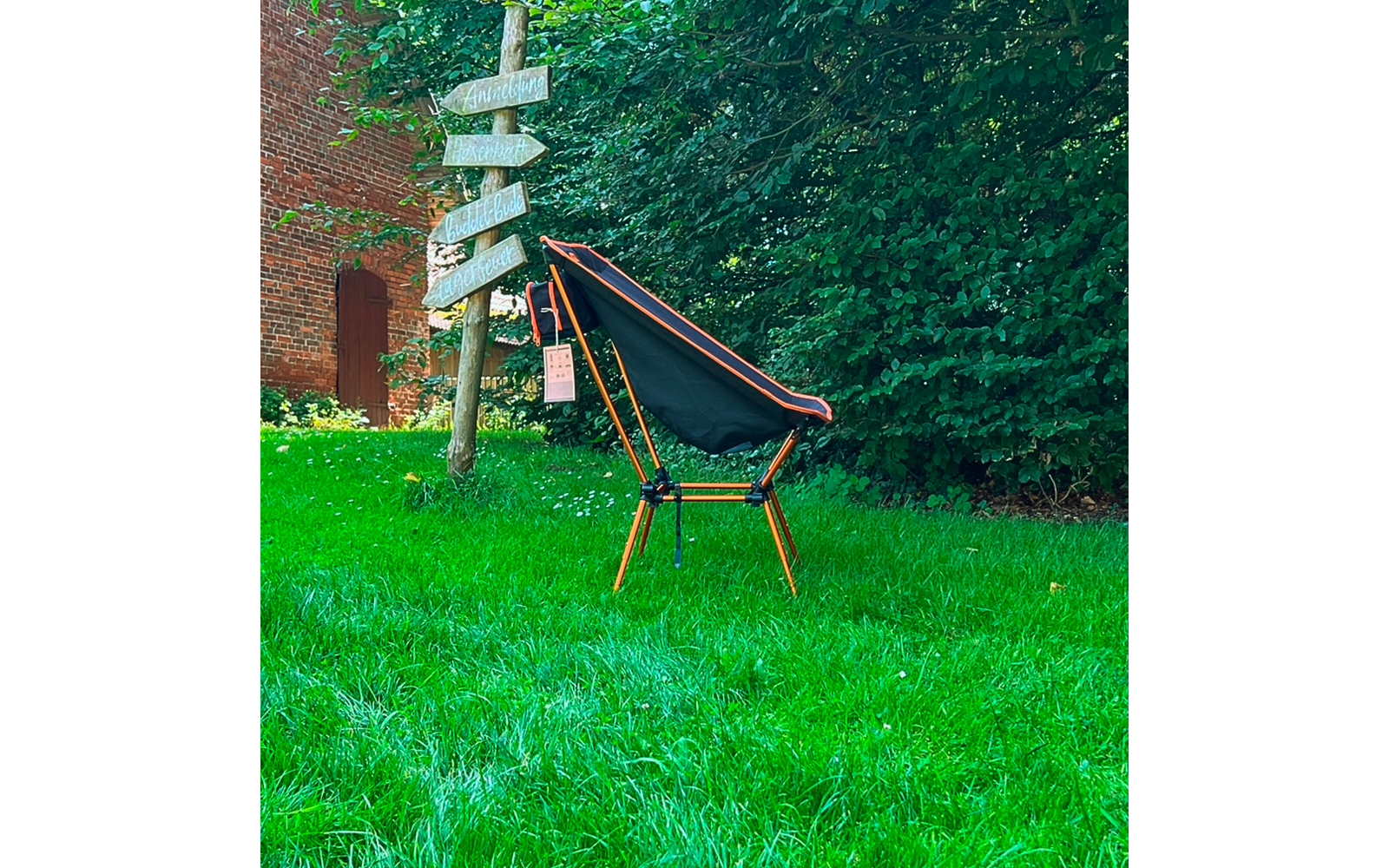 Chaise pliante noire avec armature orange sur herbe verte; à gauche un bâtiment en briques rouges et un poteau en bois avec plusieurs panneaux blancs; en arrière-plan une haie verte dense; une étiquette rose pend à la chaise