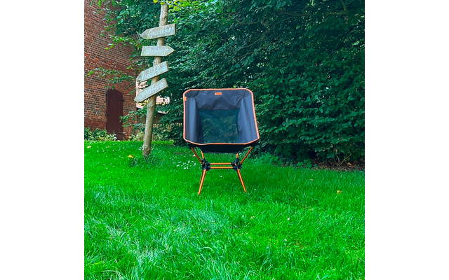 Chaise pliante avec armature orange et assise sombre sur pelouse verte ; à gauche un panneau en bois avec plusieurs flèches, en arrière-plan un mur de briques avec porte en arc et une haie
