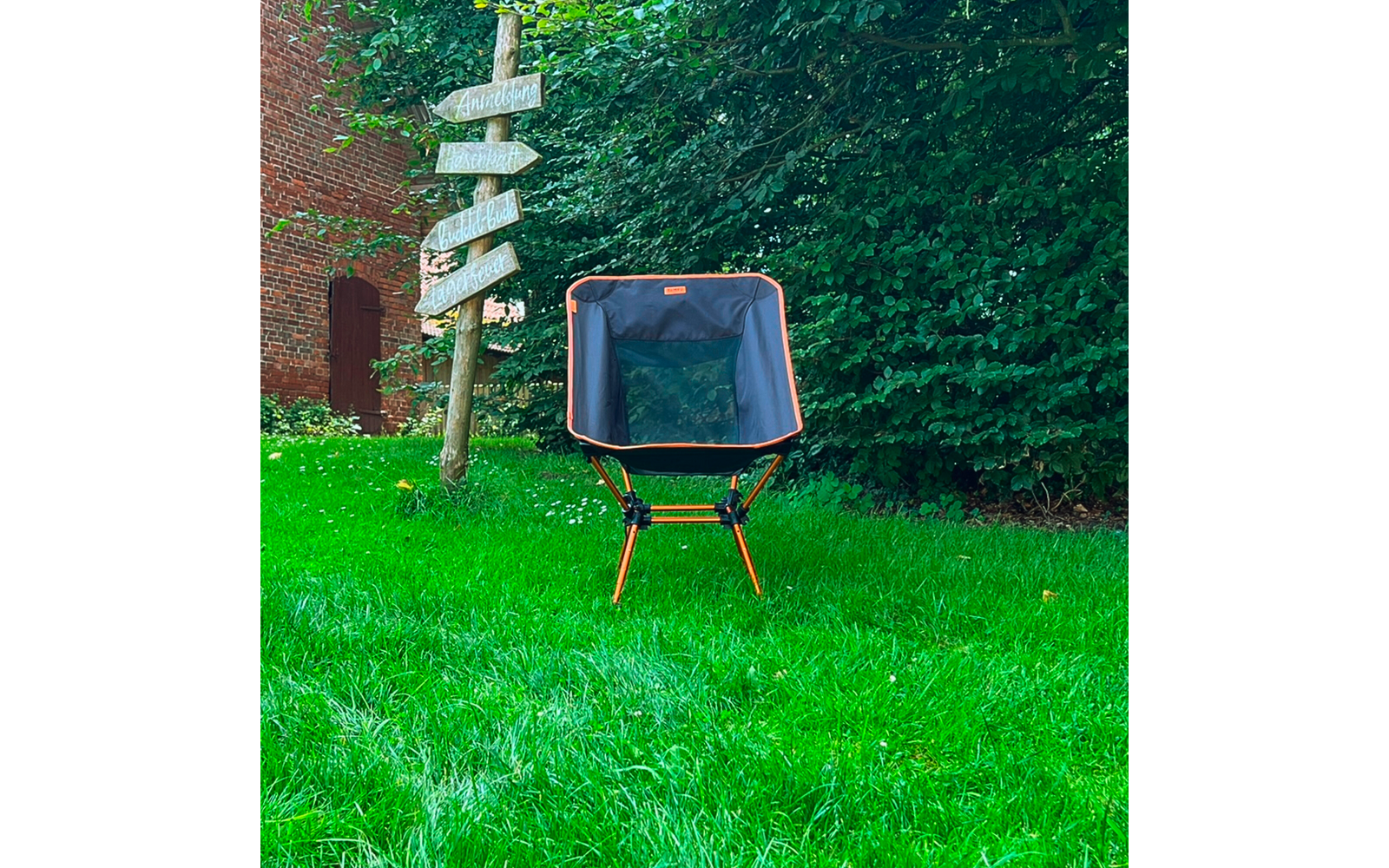Chaise pliante avec armature orange et assise sombre sur pelouse verte ; à gauche un panneau en bois avec plusieurs flèches, en arrière-plan un mur de briques avec porte en arc et une haie