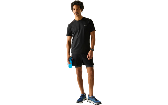 Person in schwarzem T-Shirt und schwarzen Shorts steht, hält eine blaue Trinkflasche, trägt blaue Laufschuhe und eine Armbanduhr, heller Hintergrund