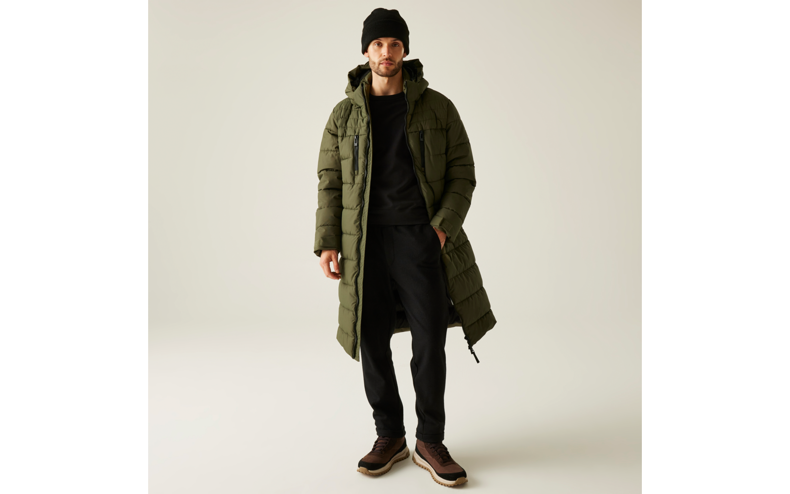 Ein Mann trägt einen langen olivgrünen Mantel, schwarzen Pullover, schwarze Hose und braune Schuhe