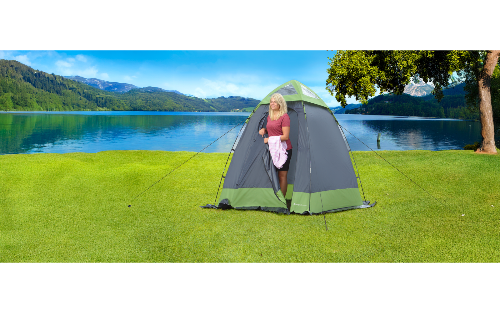 Woman standing inside a freestanding shower tent on a green lawn next to a lake with mountains in the background