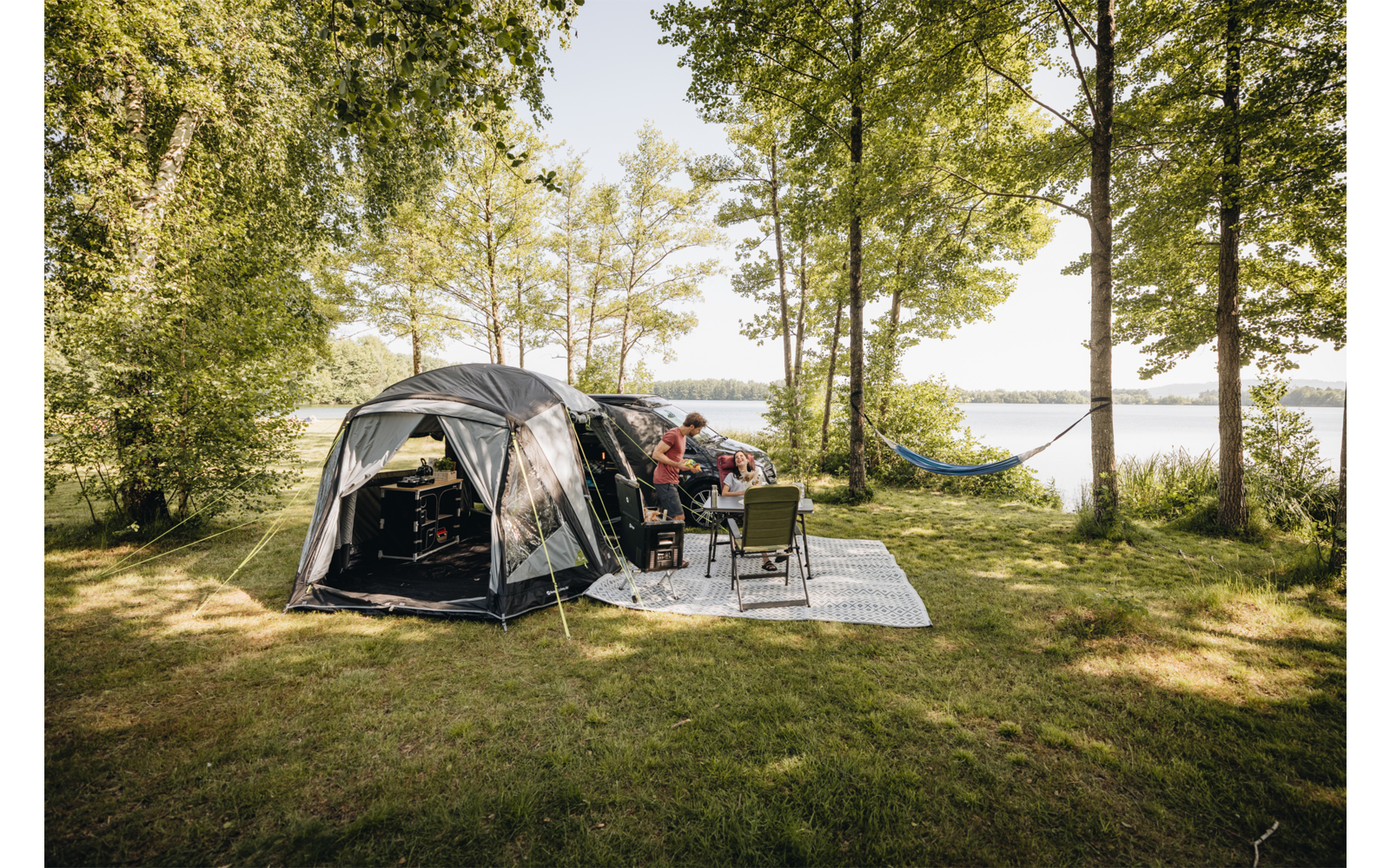 Tent on a grassy area by a lake, people sitting at a camping table, chairs and a picnic mat in front, hammock between trees, lake and trees in the background